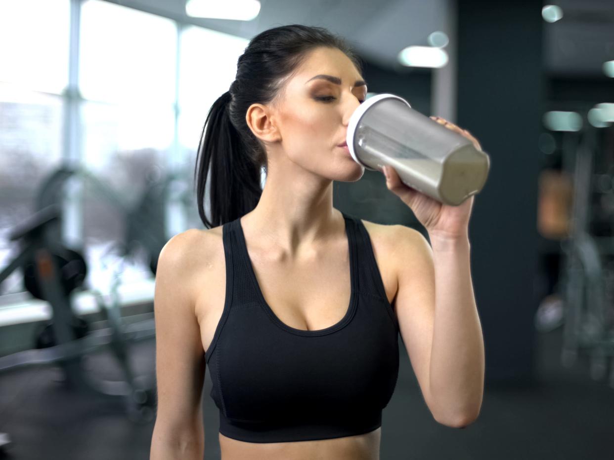 a woman at the gym drinking a protein shake, wearing a black sports bra with exercise machines in soft focus in the background