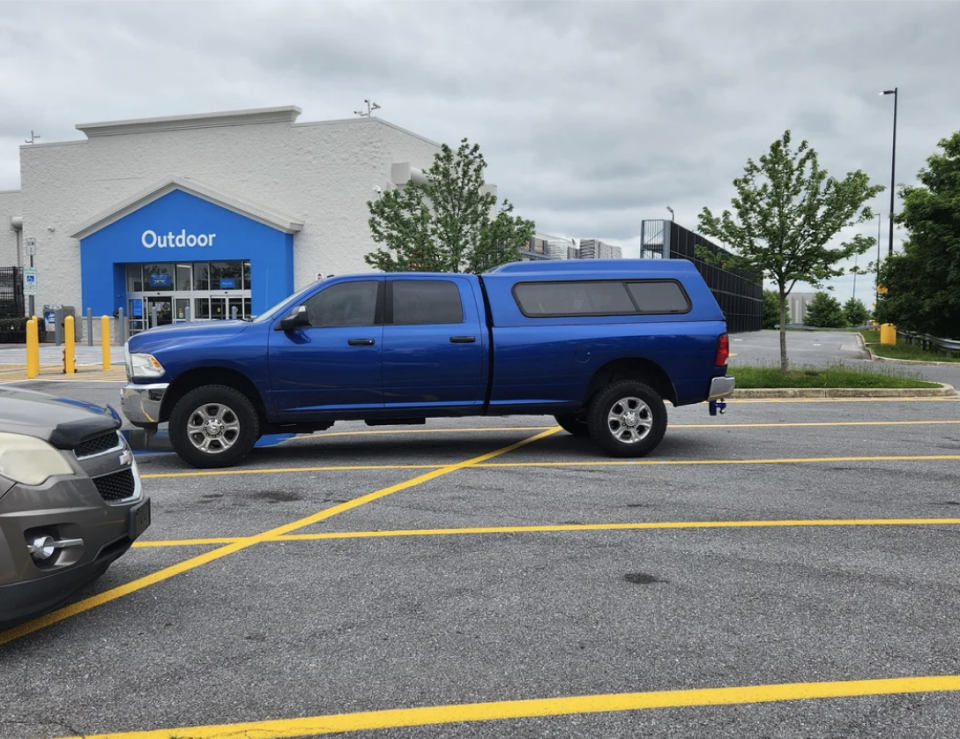 A blue pickup truck with a topper is parked in a parking lot near a store labeled "Outdoor." Trees and other cars are visible in the background