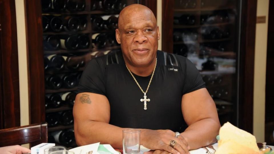 A man with dark-toned skin sits with a neutral expression at a table, a collection of wine behind him.