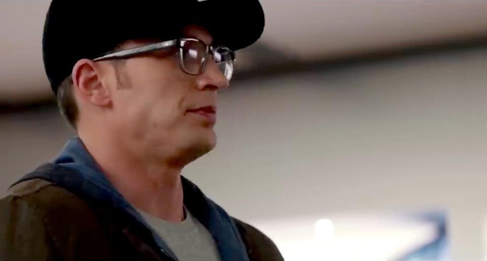 Man with glasses and cap, wearing a casual hoodie and jacket, looks to his right in an indoor setting