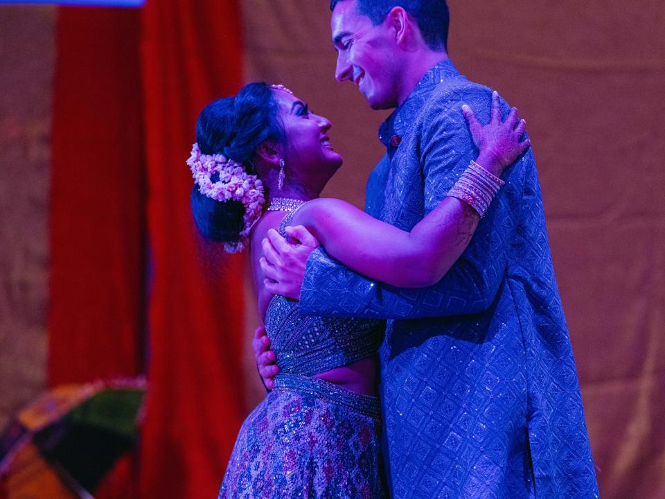 The couple are both in blue traditional Indian outfits and engaged in a dance routine. They are posed holding each other and making eye contact. The bride is wearing a white floral headpiece and white sneakers under her lehenga.