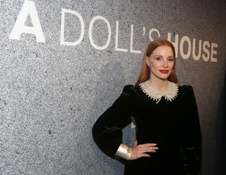 jessica chastain, wearing a black dress with white lace, puts her hand on her hip and stands in front of a stone wall with the words a doll's house on it