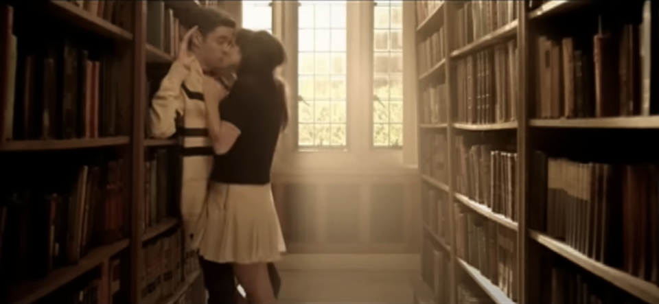 Two people are in a library aisle, with bookshelves on either side. The woman is pinning the man against the shelf passionately