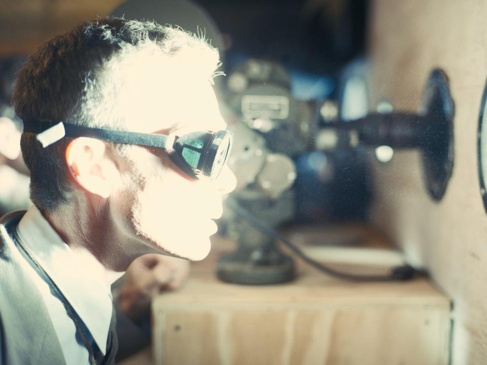 Cillian Murphy in a scene from "Oppenheimer" where Oppenheimer is seen wearing a set of googles and looking through a porthole while a bright light illuminates his face.