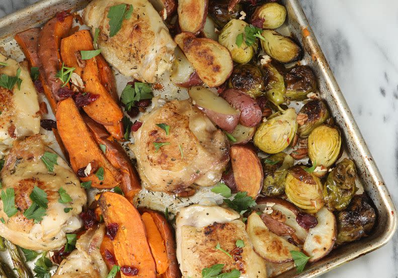 An overhead close up horizontal photograph of a freshly baked sheet pan dinner consisting of chicken thighs, acorn squash, asparagus, yams , red potatoes and Brussel sprouts. A small bowl of chopped parsley leaves sits by the pan.