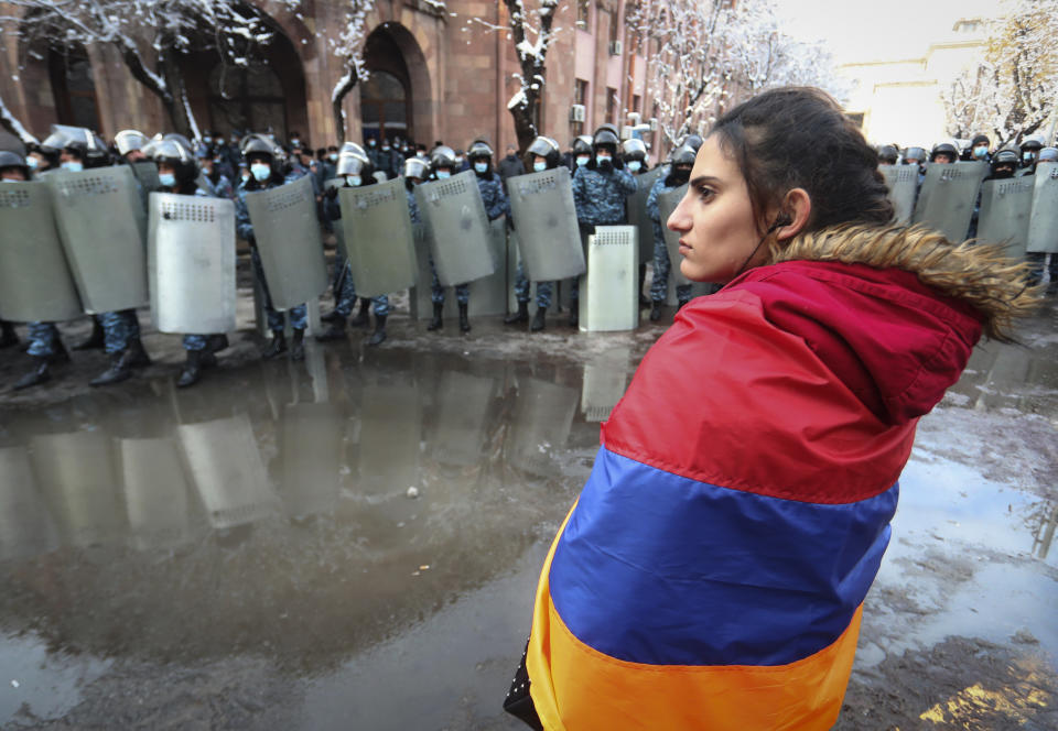 A woman wearing a state Armenian flag stands in front of a riot police line during a rally to pressure Armenian Prime Minister Nikol Pashinyan to resign over a peace deal with neighboring Azerbaijan on Republic Square in Yerevan, Armenia, Thursday, Dec. 24, 2020. Armenian opposition politicians and their supporters have been protesting for weeks, demanding the prime minister's resignation over his handling of the Nagorno-Karabakh conflict with Azerbaijan. (Vahram Baghdasaryan, Photolure via AP)
