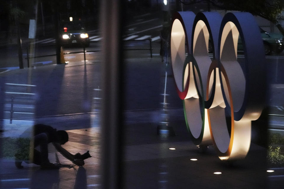 A photographer adjusting a camera to shoot the Olympic rings is reflected on a glass window near the New National Stadium Wednesday, July 22, 2020, in Tokyo. The postponed Tokyo Olympics have again reached the one-year-to-go mark. But the celebration is small this time with more questions than answers about how the Olympics can happen in the middle of a pandemic. That was before COVID-19 postponed the Olympics and pushed back the opening to July 23, 2021. (AP Photo/Eugene Hoshiko)