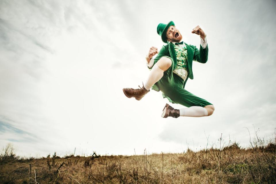 a stereotypical irish character all ready for saint patricks day jumps and dances in an open field of irish country side copy space in the sky and grass