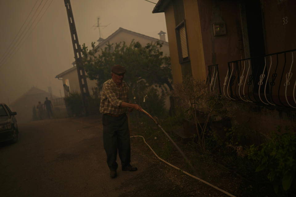 An elderly man uses a garden hose to water the outside of a house as a forest fire smoke darkens the sky in the village of Bemposta, near Ansiao, central Portugal, Wednesday, July 13, 2022. Thousands of firefighters in Portugal continue to battle fires all over the country that forced the evacuation of dozens of people from their homes. (AP Photo/Armando Franca)