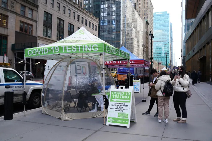 People in winter clothing gather around a temporary booth on a sidewalk in Manhattan advertising COVID-19 testing.