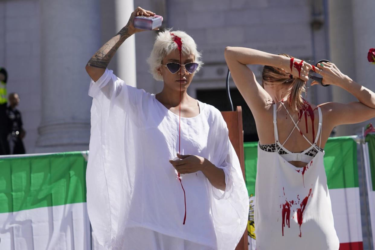 A woman with short blond hair, left, pours red liquid over her head while another woman seen from behind cuts her ponytail