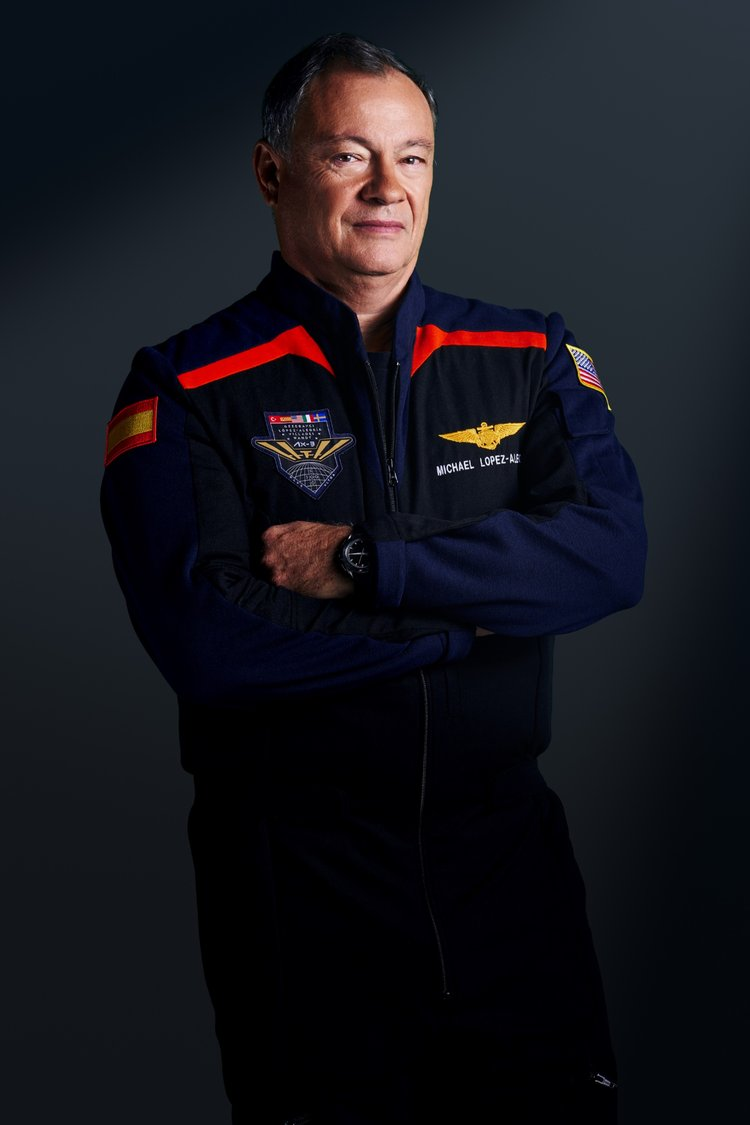 A man stands with his arms crossed, wearing a dark blue jumpsuit with wings on his shoulders with red accents.  He looks serious.