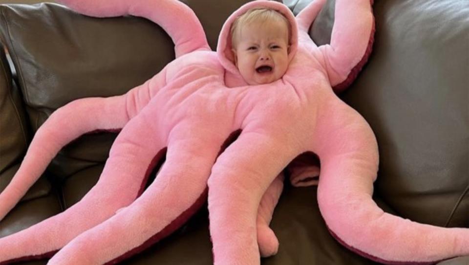 A crying baby with blonde hair dressed in a octopus costume with eight pink arms