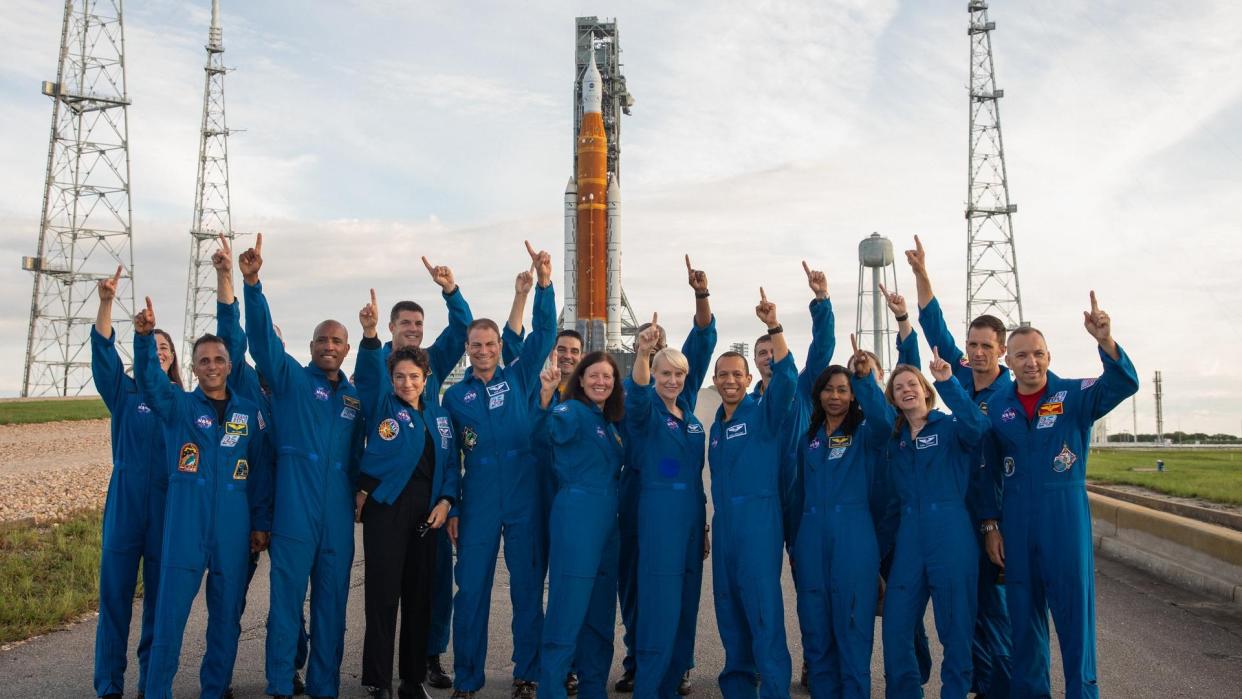  A bunch of astronauts in flight suits standing in front of a huge rocket and pointing at the sky. 