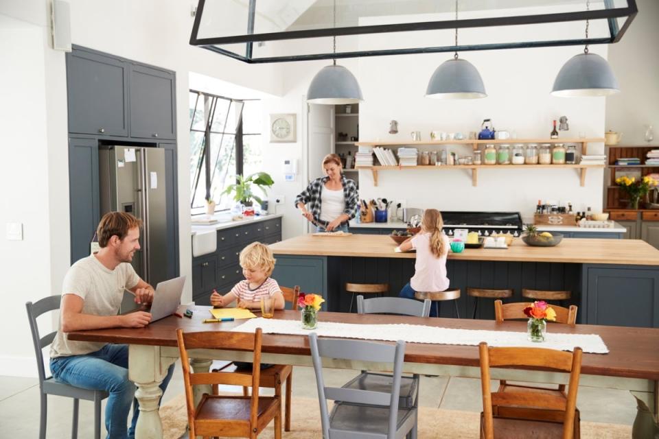 Family with two young children enjoying in a large, open plan kitchen. Mom is at kitchen island and dad is at table with laptop.