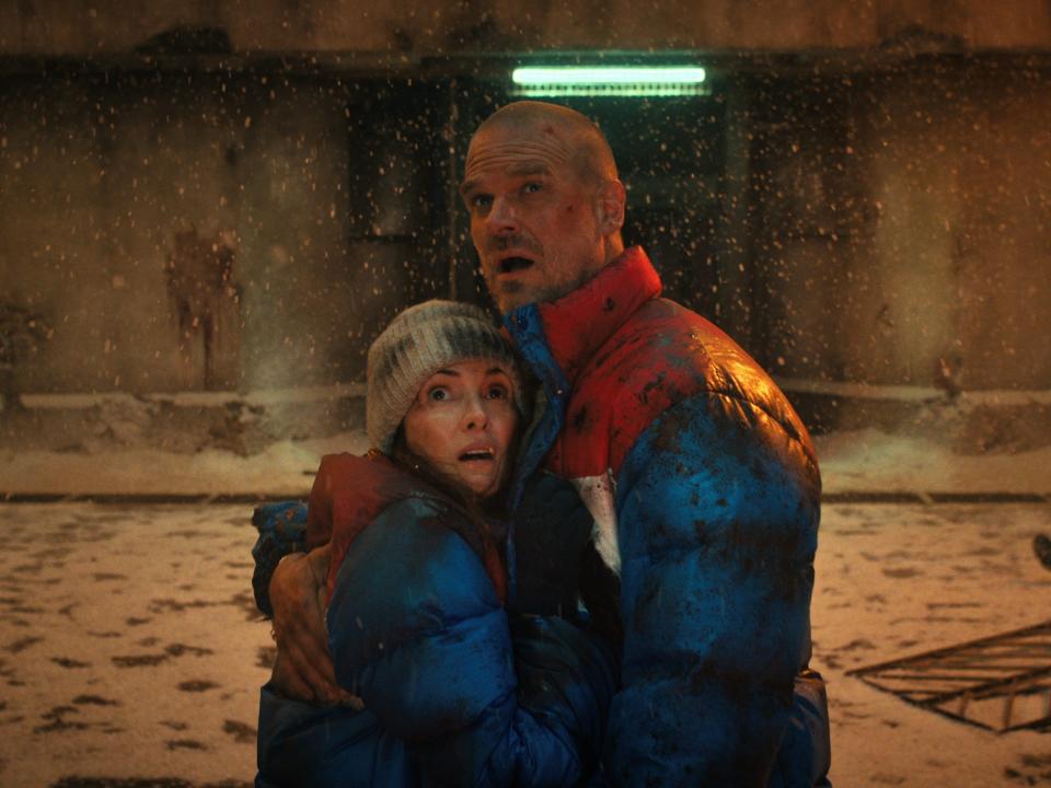 joyce byers and hopper huddled together in puffer goats, standing in the middle of a snowy, concrete-walled arena in stranger things