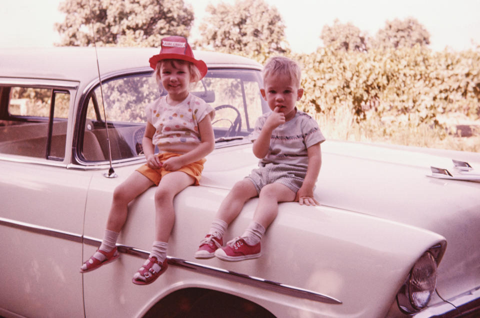 Two children, one wearing a red hat and orange shorts and the other in green striped overalls, sit on the hood of a vintage car