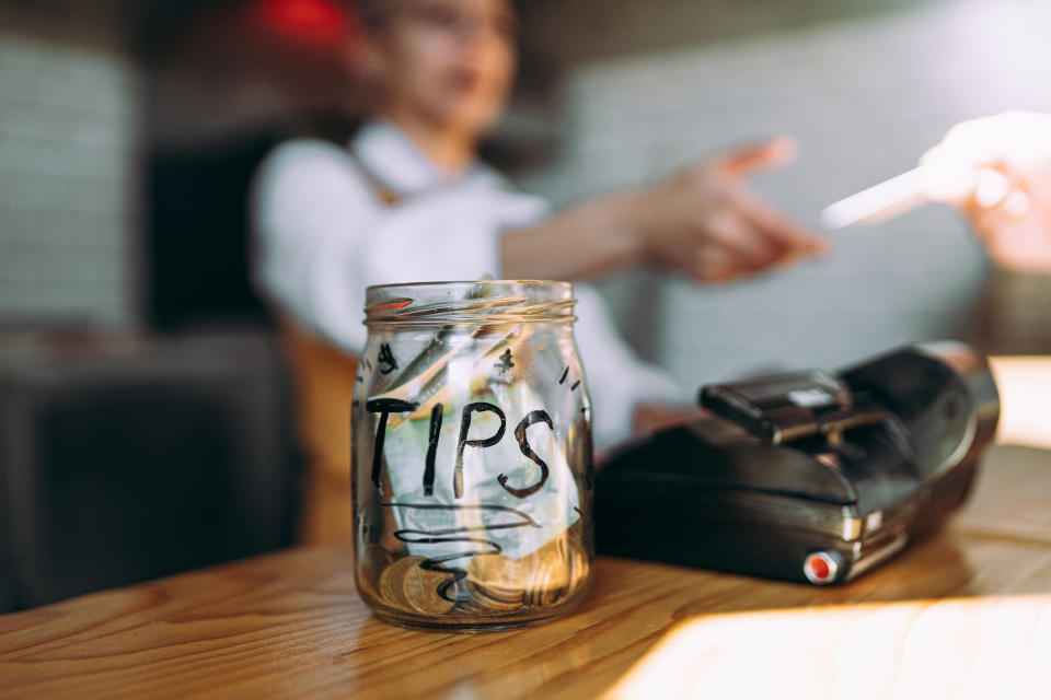 A tip jar filled with money on a counter; a person's hand is seen gesturing in the background