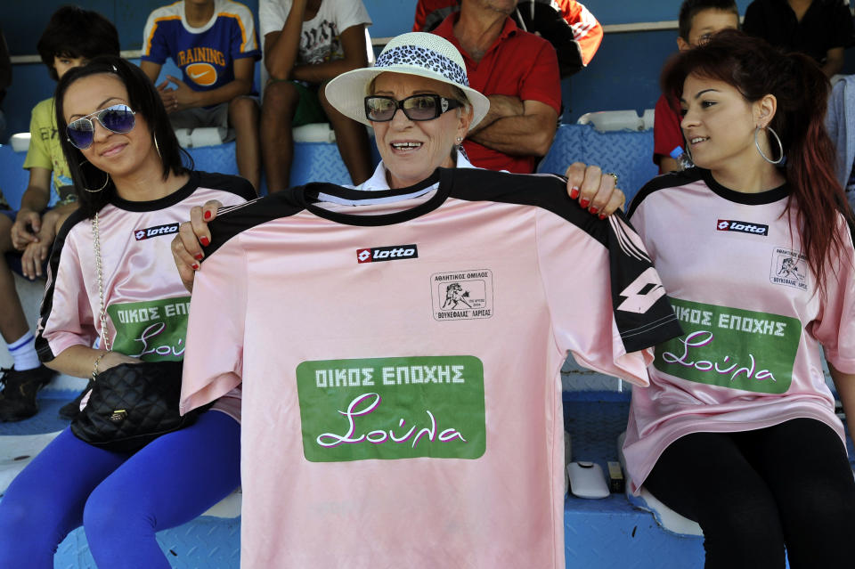 In this photo taken Sunday, Oct. 7, 2012, brothel owner Soula Alevridou, centre, the benefactor of the Voukefalas amateur soccer team, holds up their new jersey for photographers, during a local championship match, in the city of Larissa, central Greece. A cash-strapped Greek soccer team has found a new way to pay the bills, with help from the world’s oldest profession. Players are wearing bright pink practice jerseys emblazoned with the logos of the Villa Erotica and Soula’s House of History, a pair of pastel-colored bordellos recruited to sponsor the team after drastic government spending cuts left the country's sports organizations facing ruin. One team took on a deal with a local funeral home and others have wooed kebab shops, a jam factory, and producers of Greece’s trademark feta cheese. But the small amateur Voukefalas club which includes students, a bartender, waiters and pizza delivery drivers is getting the most attention for its flamboyant sponsors. Logo on T-shirt translates to 'Soula's House of History'. (AP Photo/Nikolas Giakoumidis)