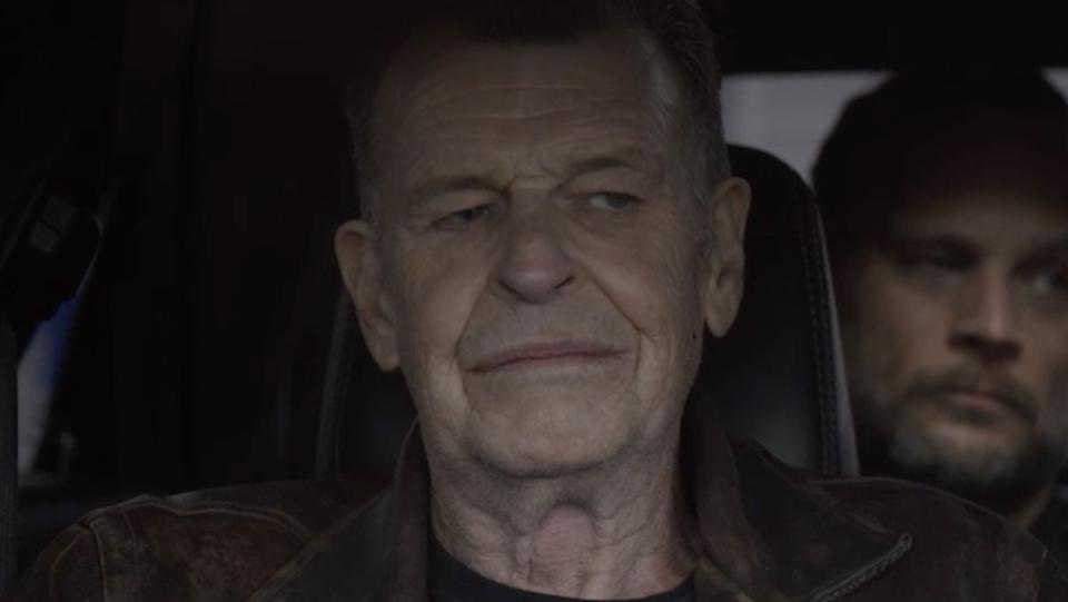 An older man in a brown leather coat sits in the passenger seat of a car with another man behind him over his shoulder