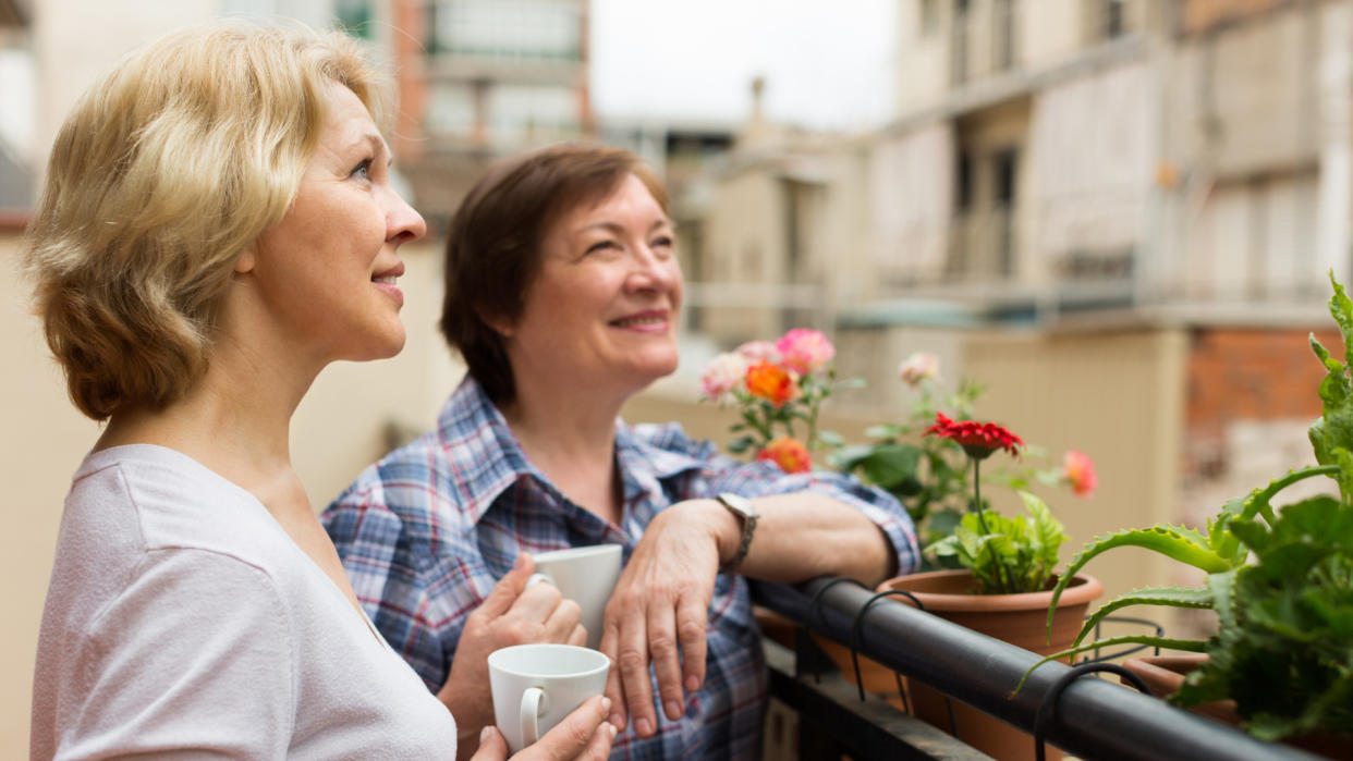 Smiling aged female friends relaxing with cup of coffee on balcony.