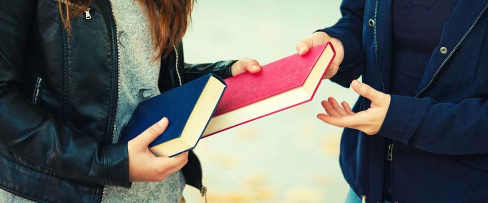 Close up outdoors portrait of two women students exchanging books. Knowledge sharing between people. Give to read to a friend.