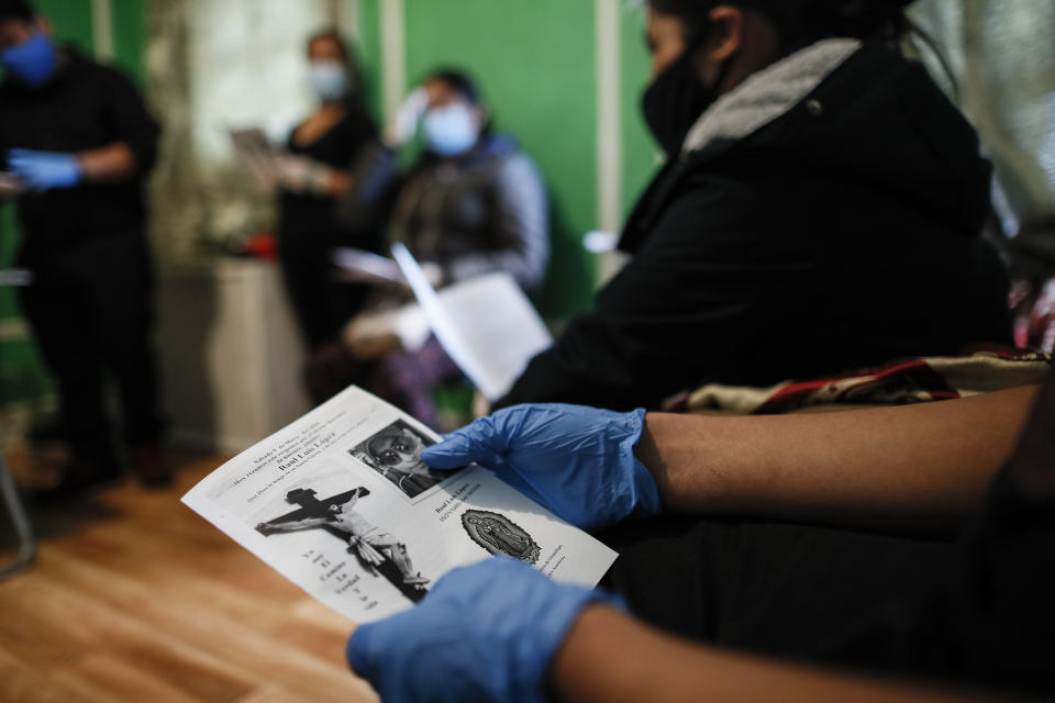 A family member holds a mass card as he wears personal protective equipment while the Rev. Fabian Arias performs an in-home service beside the remains of Raul Luis Lopez who died from COVID-19 the previous month, Saturday, May 9, 2020, in the Corona neighborhood of the Queens borough of New York. (AP Photo/John Minchillo)