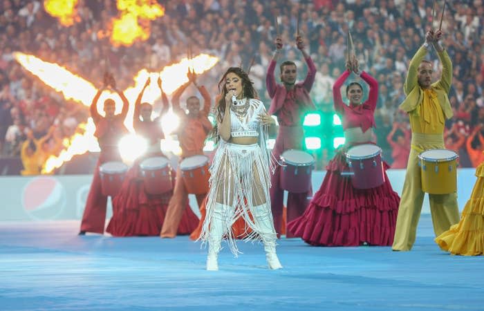 Camila performing on a soccer field, surrounded by drummers wearing colorful outfits