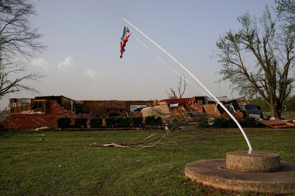 A flagpole bent to a 45-degree angle to the ground, with a huge branch on the lawn beside it and a flattened ranch house in the background.