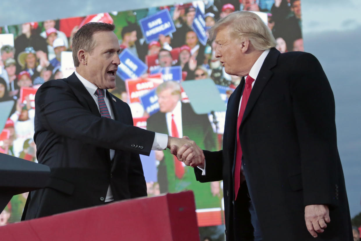 Ted Budd and Donald Trump exchange an enthusiastic handshake in front of a poster showing Trump at a rally in front of a cheering crowd.