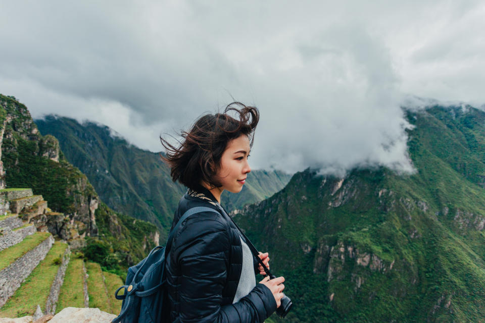 A woman stands on a mountainous landscape, holding a camera and looking off into the distance. Clouds hover over the green peaks in the background