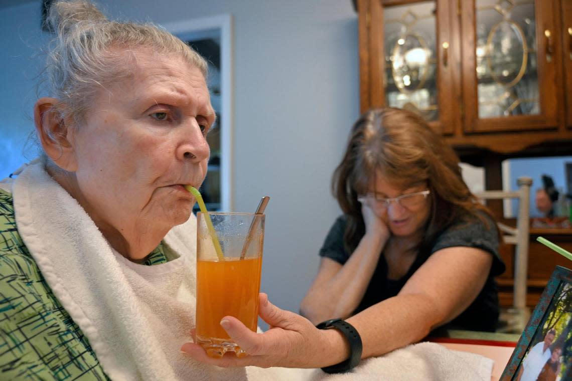 Caregiver Carol Dowell checks her messages as she holds a glass of Metamucil for Ellen DeFoe, her former mother-in-law, who has late-stage Alzheimer’s disease. “She can do nothing on her own,” Dowell said of DeFoe, who is immobile and mostly non-verbal.