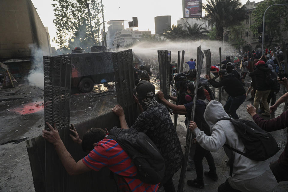 In this Oct. 29, 2019 photo, anti-government protesters shield themselves from a police water cannon in Santiago, Chile. Demonstrations that began with youth protests over a subway fare hike transformed into a leaderless national movement demanding greater equality and better public services in a country long seen as an economic success story. (AP Photo/Esteban Felix)