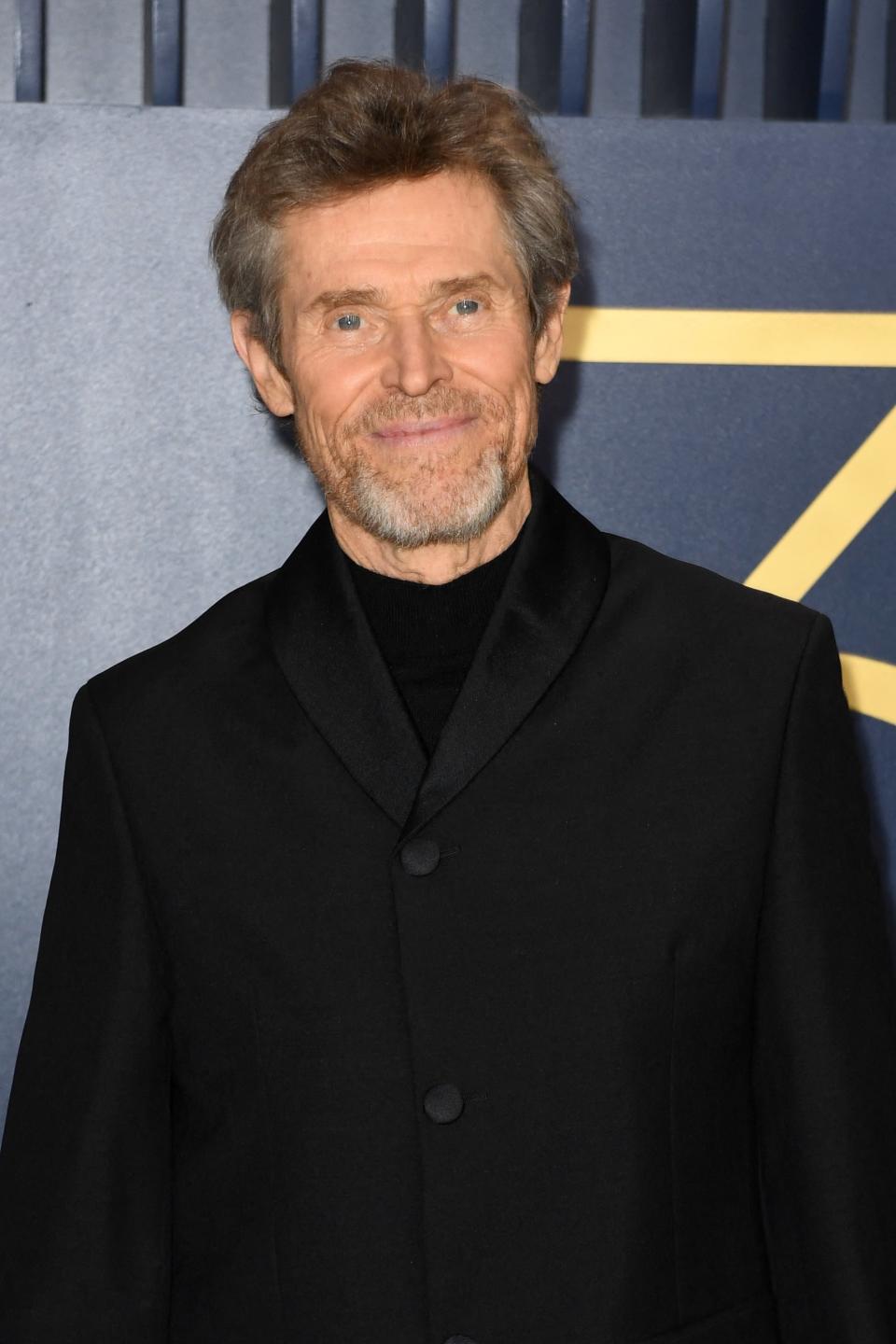 Willem Dafoe smiling in a simple suit on the red carpet