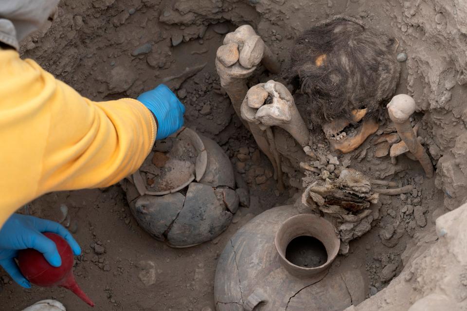 A mummy with hair on its head sits in a shallow grave.