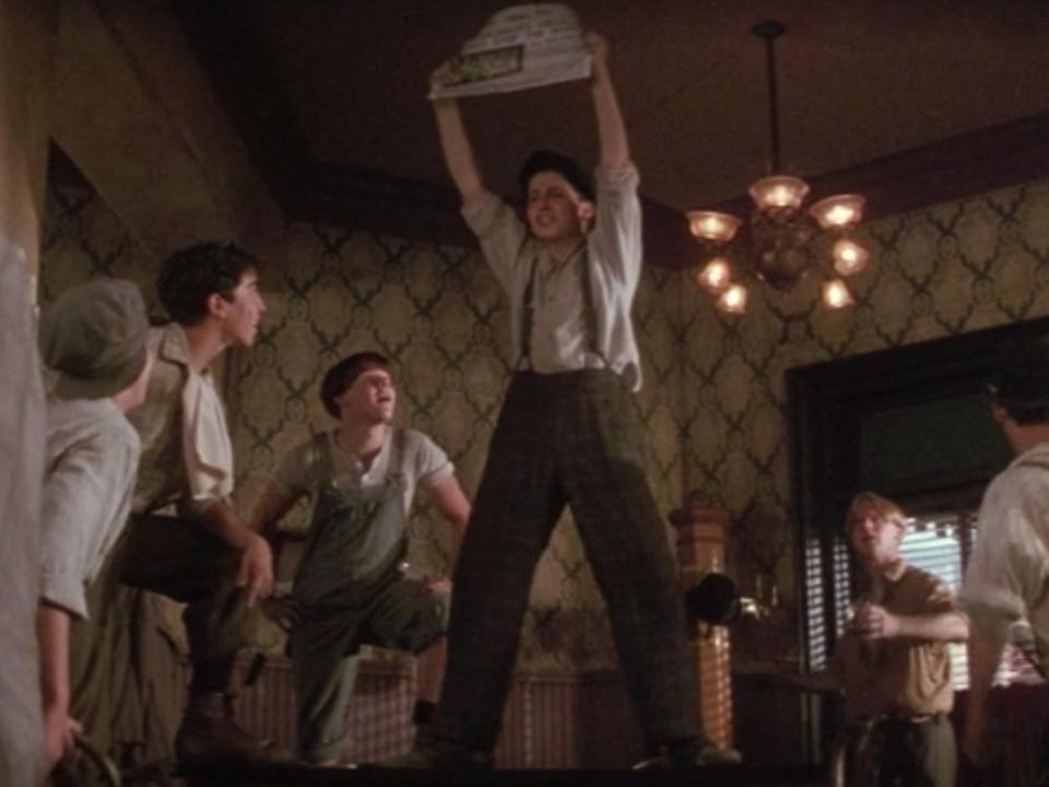 a newsie on the table holding a newspaper in Newsies king of new york