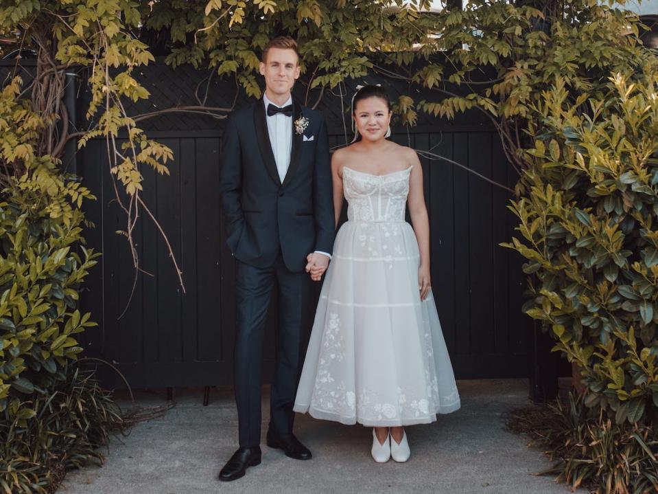 A bride and groom hold hands in front of greenery.