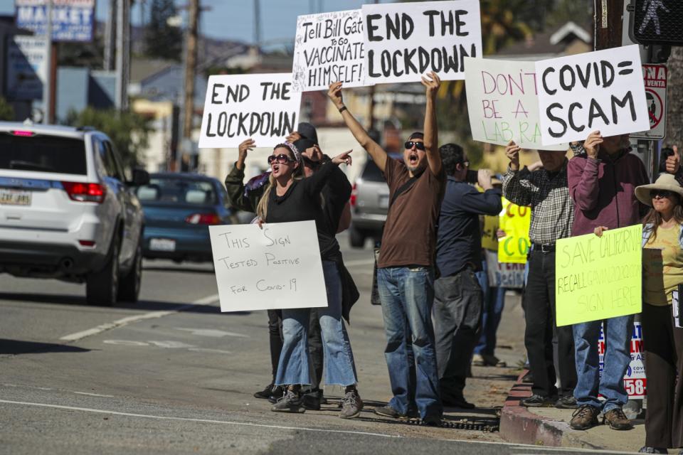 People yell on a street corner. Signs include "This sign tested positive for COVID-19" and "COVID = Scam."