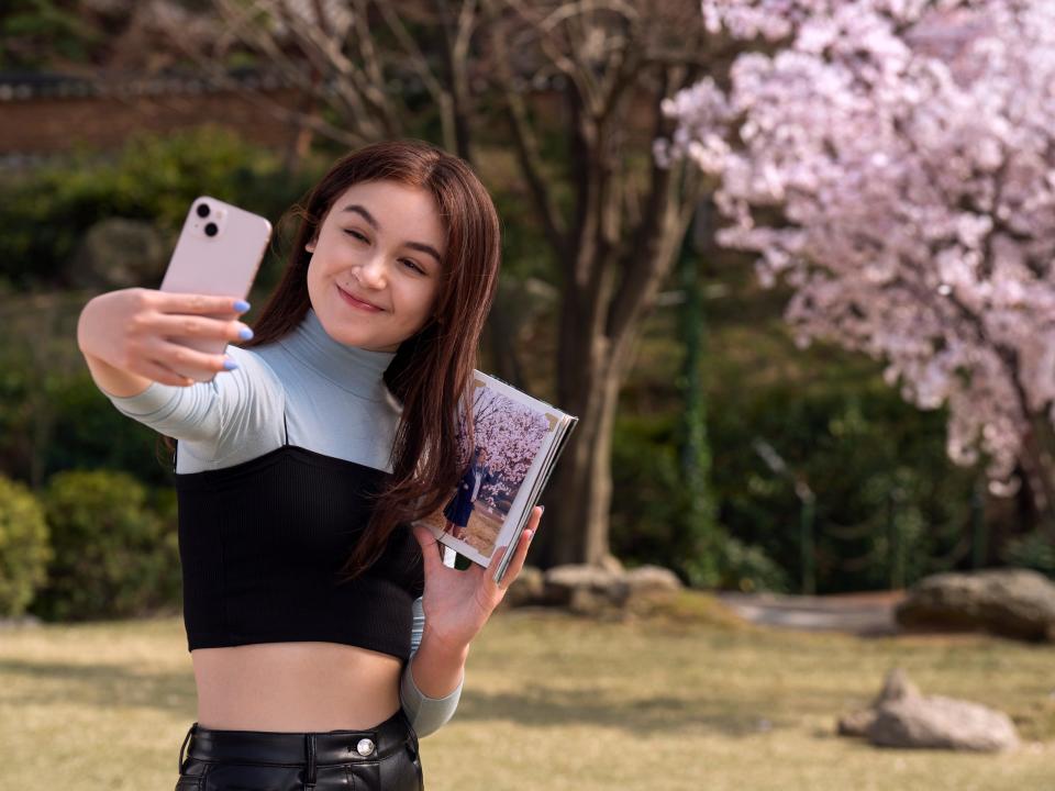 anna cathcart as kitty in xo kitty. she's a teenage girl wearing a blue turtleneck and cropped black tee, holding a photo of her mother in front of a cherry blossom tree. kitty snaps a selfie of herself, posing in front of the same tree.