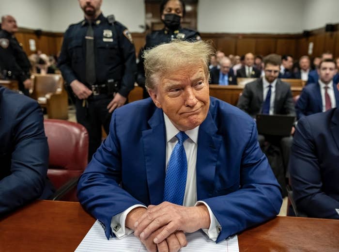 Donald Trump, seated in a courtroom, wearing a suit and patterned tie, with police officers and lawyers in the background