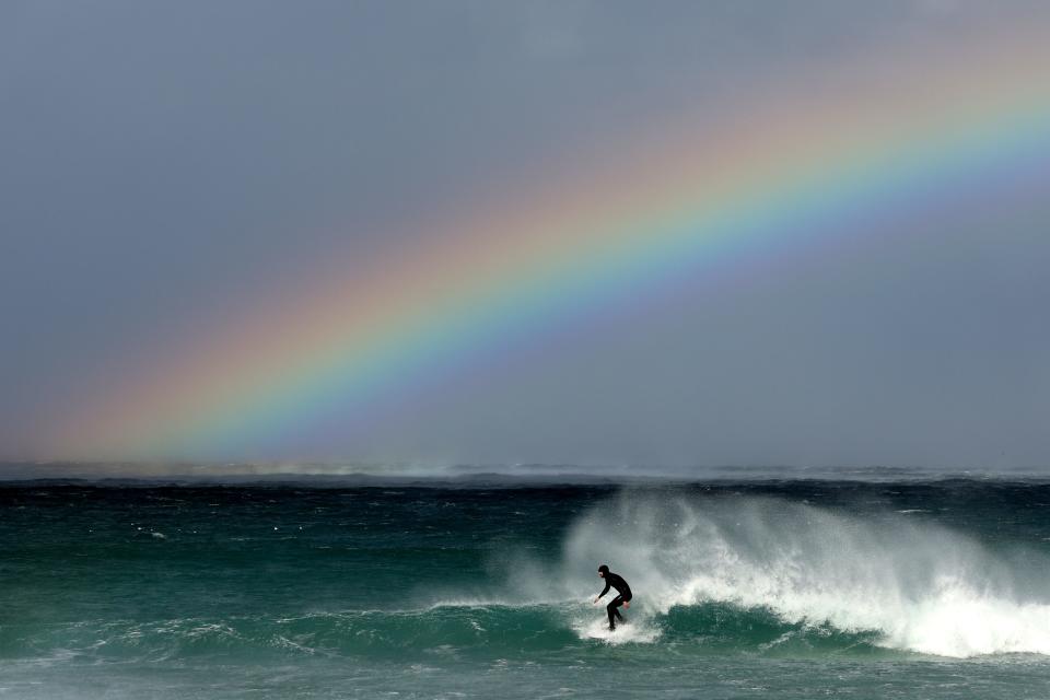 PEBBLE BEACH, CALIFORNIA - FEBRUARY 04: A surfer surfs under a rainbow near the sight of the AT&T Pebble Beach Pro-Am at on February 04, 2024 in Pebble Beach, California. Sunday's final round was postponed due to inclement weather.