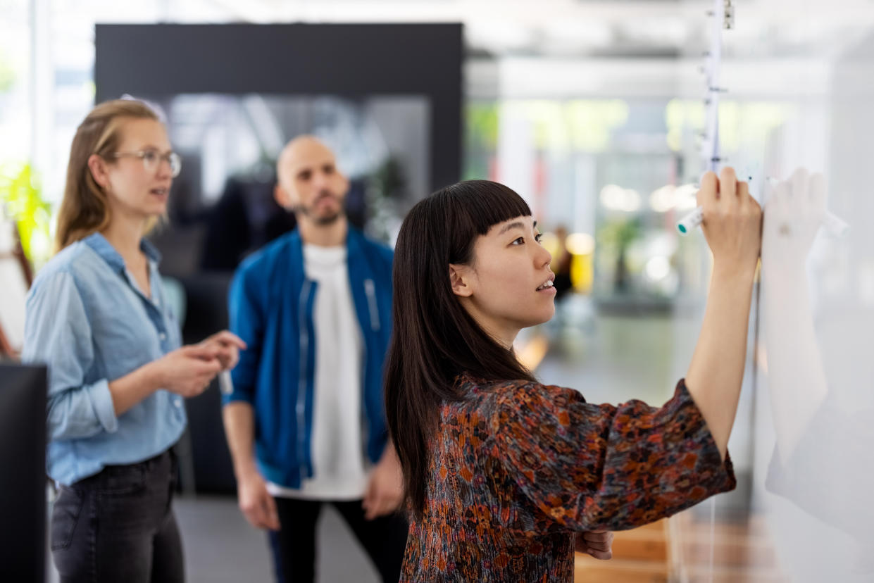 Young businesswoman explaining a business plan to colleague in office. Woman writing on the whiteboard during presentation in hybrid office space.