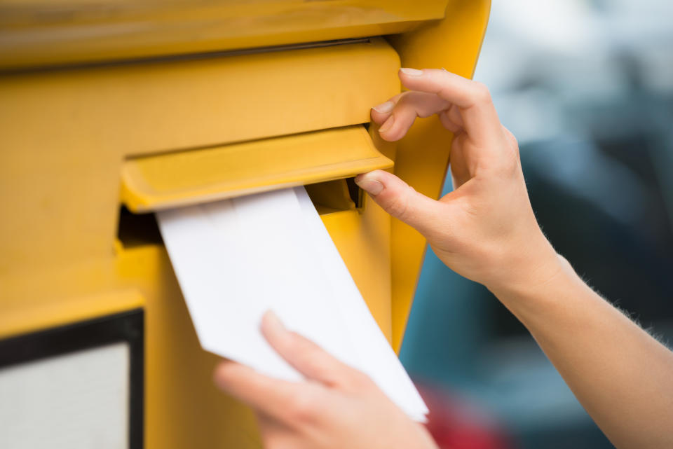 A person is mailing an envelope in a yellow mailbox. The image focuses on the hands as they insert the envelope