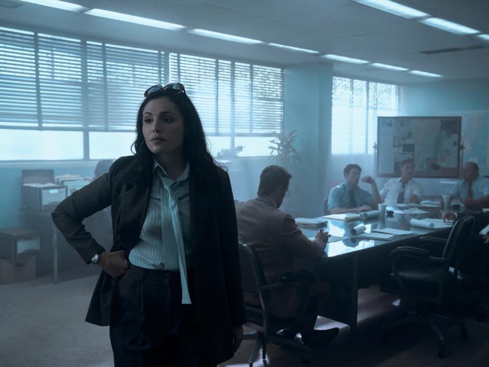 juliana aidén martinez as june in griselda, wearing a pantsuit and standing in front of a long table of men