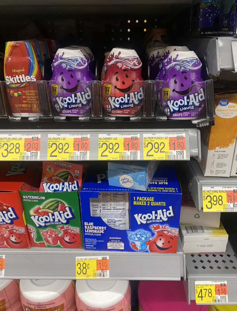 Shelf with assorted Kool-Aid drink mixes, Skittles candy, and other beverages. Prices and product labels are visible below each item