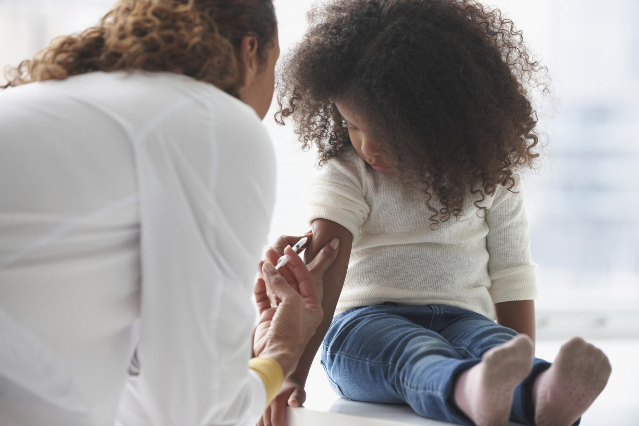 A health care worker gives a shot into the upper arm of a young girl sitting on a table and looking at the needle.