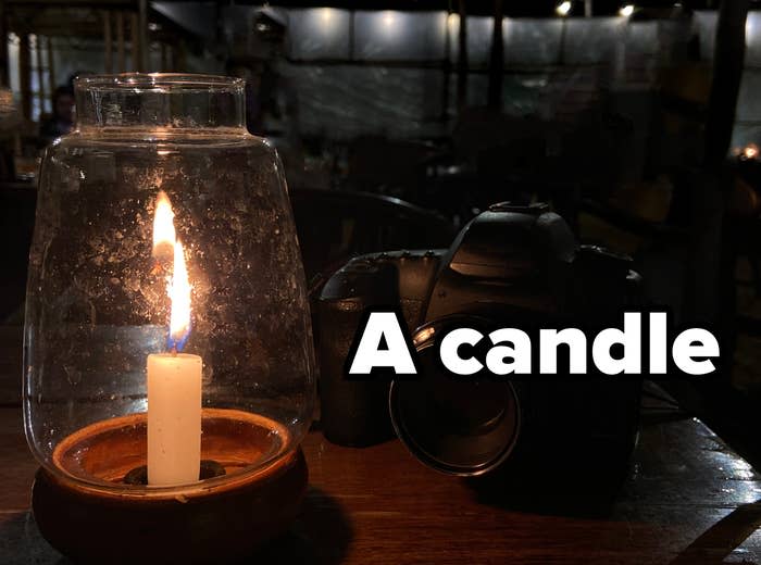 A lit candle in a glass holder and a camera on a wooden table in a dimly lit room