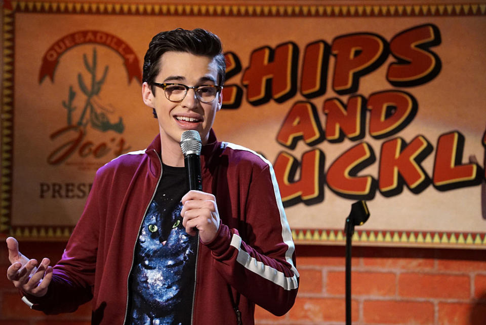 Joey in a scene on stage holding a microphone with graphic tee and jacket, in front of a sign reading "Chips and Chuckles"