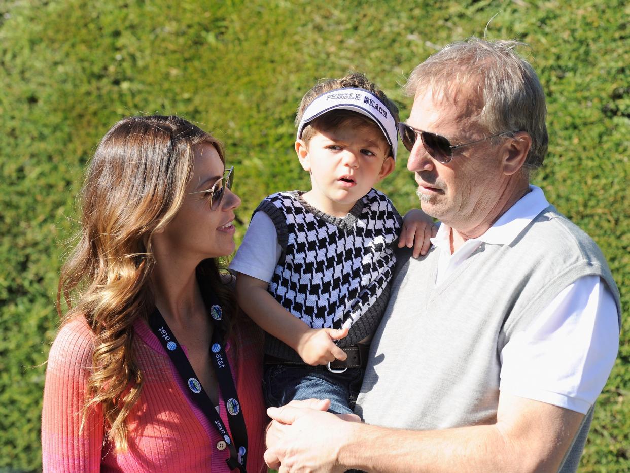 Kevin Costner and Christine Baumgartner standing together in front of a hedge while holding one of their children.