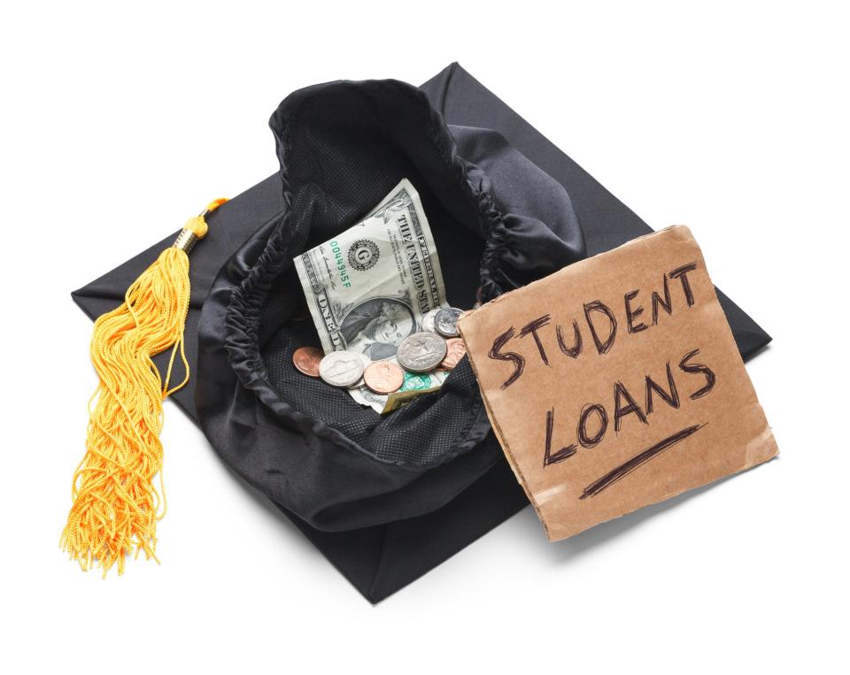 Upside-down graduation cap with dollar bill and coins in it, next to brown sign that says student loans